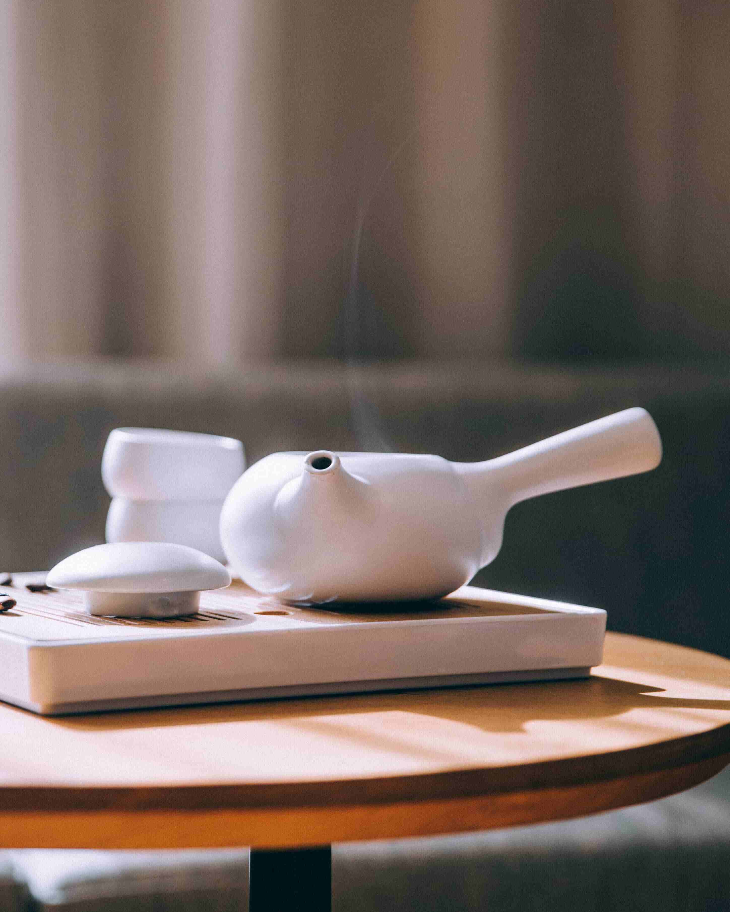 A Chinese tea pot sitting steaming on top of a book which lays on top of a round wooden side table. 